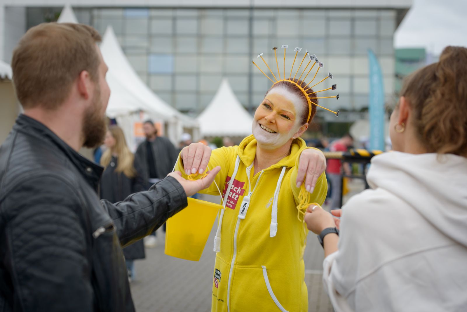 Maskottchen Sunna übergibt Sonnen-Pfäden an zwei Besucher*innen